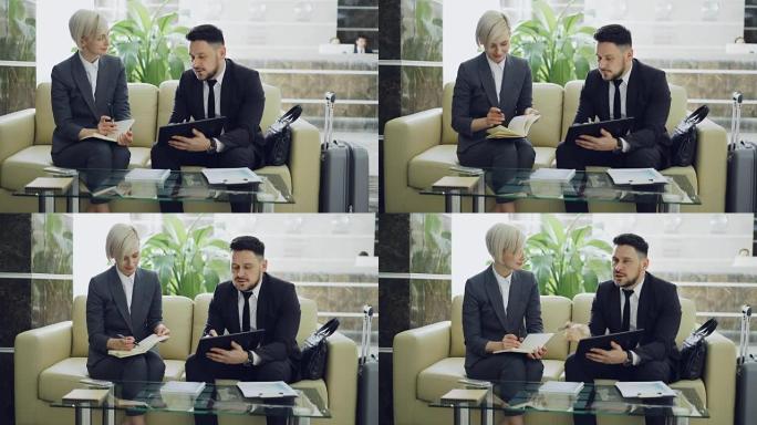 两名男女商业同事使用数字平板电脑，记事本并在酒店大堂讨论项目，同时坐在沙发上等待接待处的入住