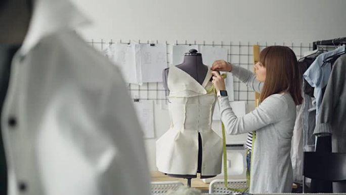 漂亮的年轻女子正在用别针调整剪裁假人的衣服，同时在轻型裁缝店的新女装上工作。创造新的服装概念。