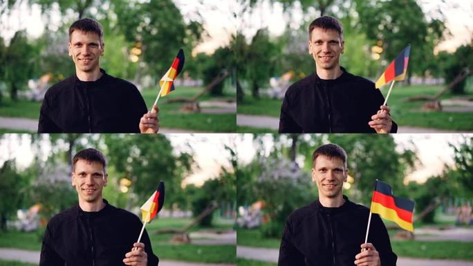 慢动作肖像欢快的德国球迷挥舞官方旗帜，看着镜头，而站在伟大的现代公园美丽的树木和草坪。