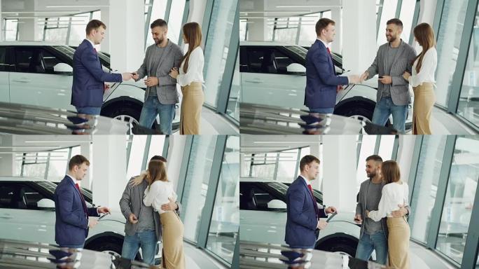 英俊的男性购车者正在从友好的推销员那里获得密钥卡，并与他握手，然后站在汽车旁边亲吻并拥抱他迷人的女友