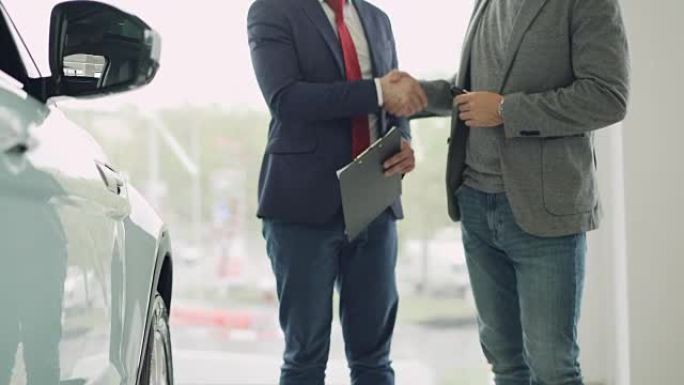 汽车陈列室的员工将汽车钥匙交给年轻的男性买家并握手，经销商拿着文件并打手势。买卖运输概念。