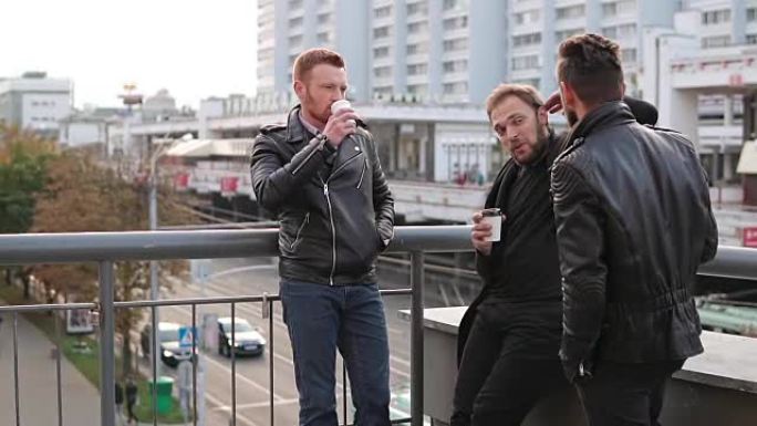 三个留着胡须的年轻英俊男子在桥栏杆上交谈。帅哥说话，喝咖啡。慢mo