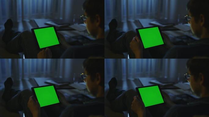 男人在夜间躺在沙发上，并在景观模式下使用带有绿屏的平板电脑