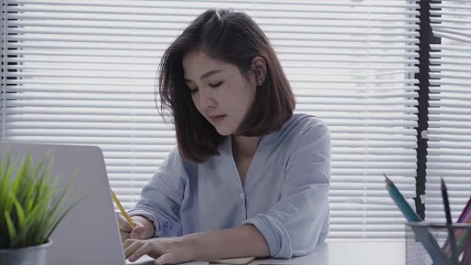 美丽的年轻微笑的亚洲女人坐在家里的客厅里在笔记本电脑上工作。在家庭办公室工作的亚洲女商人。在家享受时