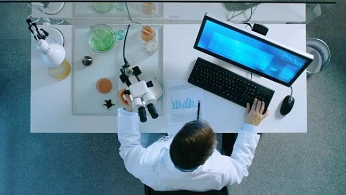 在个人计算机上工作的科学家的俯视图，观察显微镜并在带有图表的文档上写下结果。