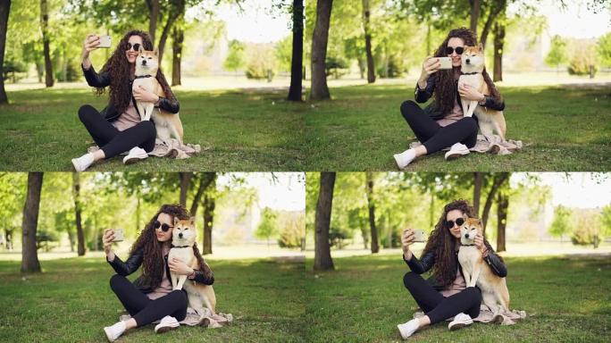 开朗的博主宠物主人正在用智能手机和她的狗自拍，人类和动物坐在公园的草坪上摆姿势，女人在爱抚和亲吻狗。