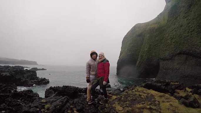 年轻的旅行夫妇穿着雨衣站在海边，享受着旅行。游客男人和女人