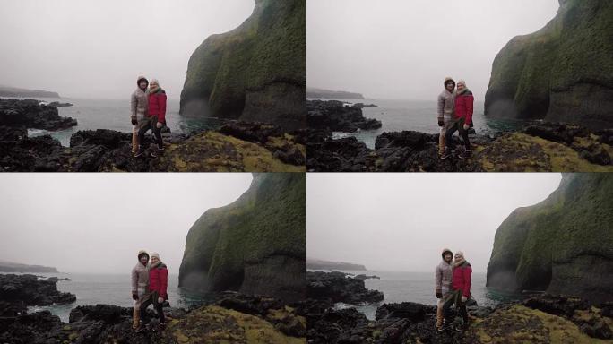 年轻的旅行夫妇穿着雨衣站在海边，享受着旅行。游客男人和女人