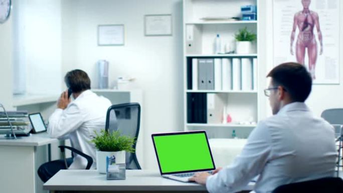 男医生在绿屏笔记本电脑上工作，他的助手在后台工作。办公室现代、宽敞、轻便。