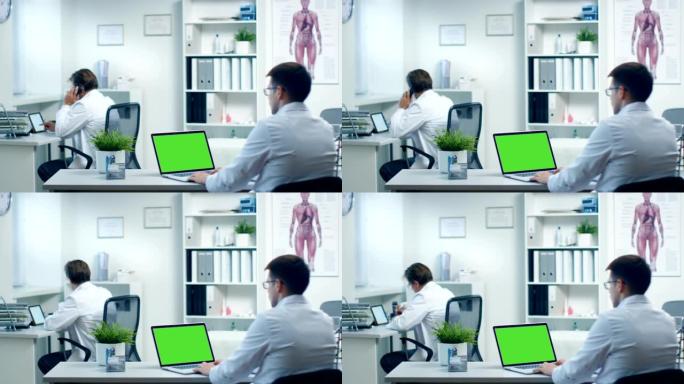 男医生在绿屏笔记本电脑上工作，他的助手在后台工作。办公室现代、宽敞、轻便。