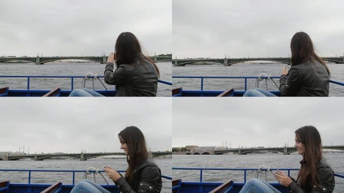 年轻的黑发女人在河巴士上观光。在缓慢的圣彼得堡的一座桥梁的智能手机上拍照
