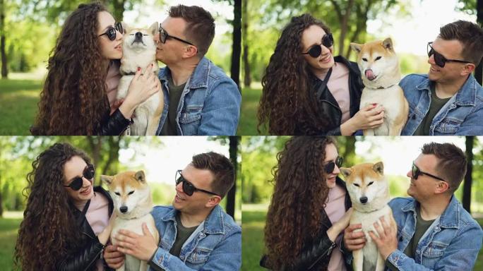 快乐的情侣慢动作亲吻美丽的狗，笑着坐在公园的草地上聊天，享受夏天。爱动物、关系和休闲概念。