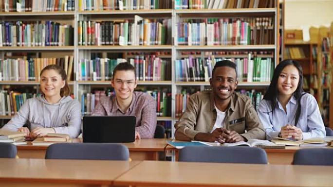 四个多民族学生的肖像坐在宽敞的大图书馆的长桌子上，成堆的书看着相机并积极微笑
