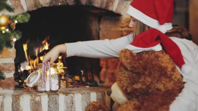 4k女孩和泰迪熊戴着圣诞老人帽子，在壁炉旁吃棉花糖，实时