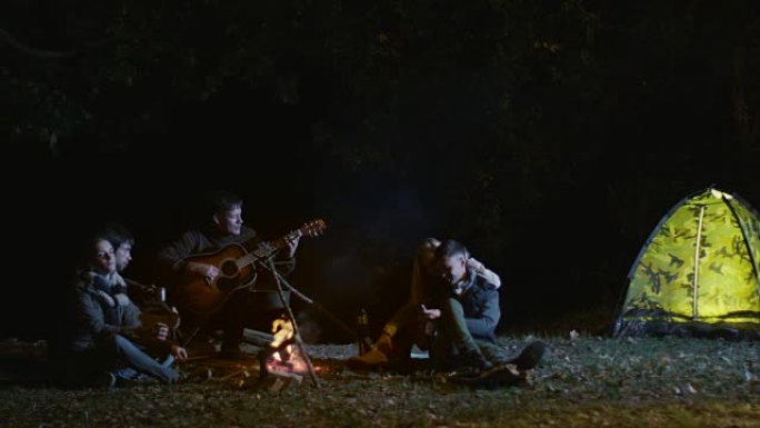 晚上，一群朋友在听，男人在篝火旁弹吉他。