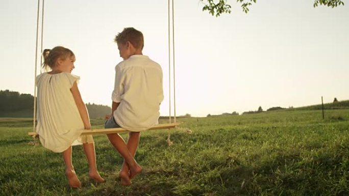 慢动作特写激动的年轻男孩和女孩在日落时在木头秋千上聊天