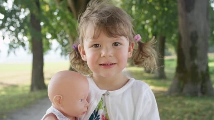 慢动作: 微笑的小女孩站在公园里抱着她的塑料玩具娃娃。