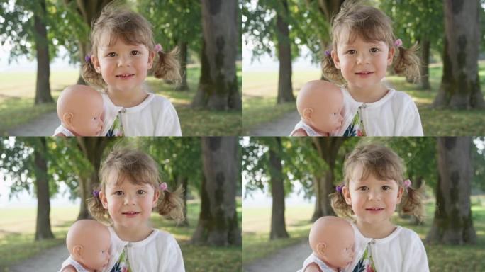慢动作: 微笑的小女孩站在公园里抱着她的塑料玩具娃娃。