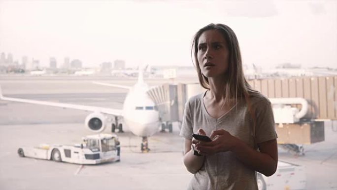 年轻的高加索女性旅行者使用智能手机，在听机场窗口附近的公告后感到沮丧