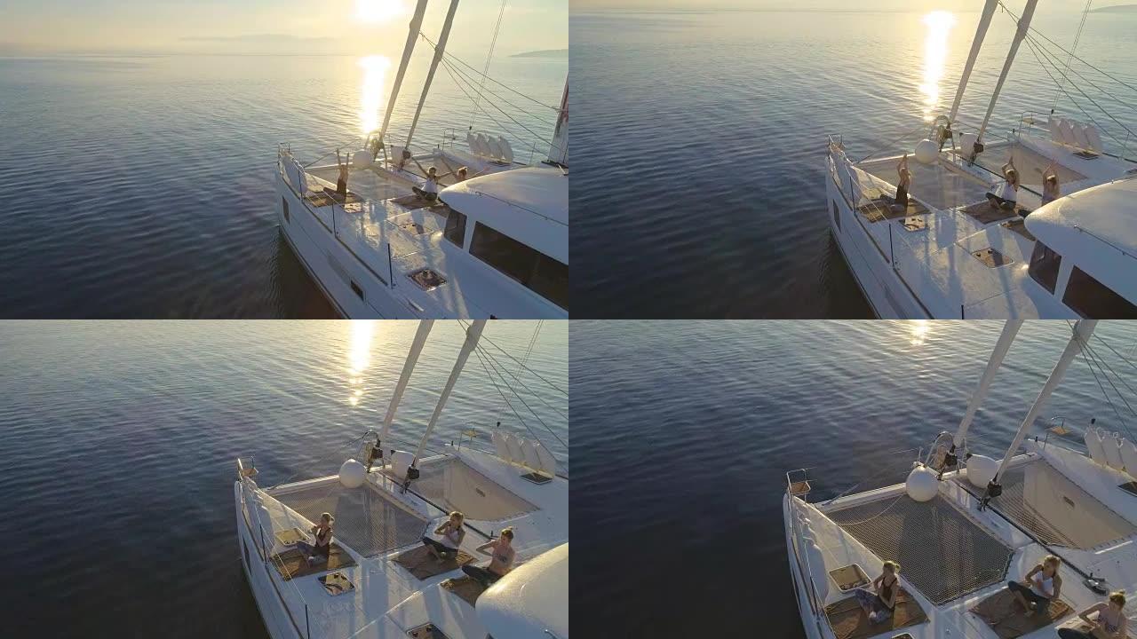 三个美丽健康的女人在一艘白色帆船双体船上做早晨瑜伽的鸟瞰图。平静的大海，太阳升起，平静的大海。