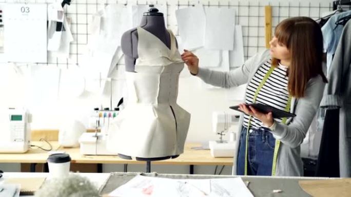 年轻的时装设计企业家正在使用平板电脑，同时检查固定在裁缝假人上的服装。服装制造概念中的现代技术。