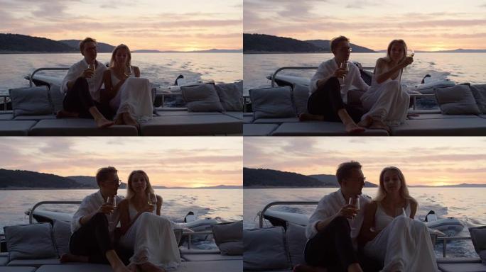 年轻夫妇拥抱，在移动的游艇船尾喝香槟。他们度过了浪漫的夜晚。在有小村庄的背景岛上。