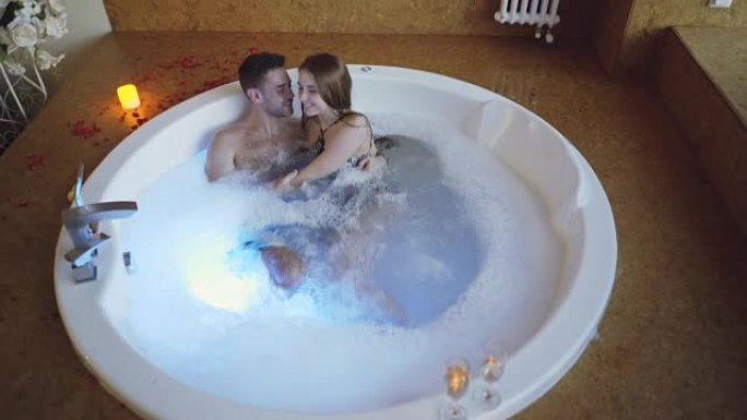 穿着泳衣的年轻人在日间水疗中心用泡沫在热水浴缸里聊天、拥抱和接吻的高角度视角。浪漫关系、激情和健康概