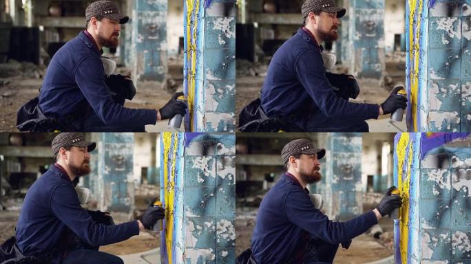严肃的成年男子涂鸦艺术家戴着皮手套在受损的空工业建筑内的柱子上绘画并蹲着。创造力和人的概念。
