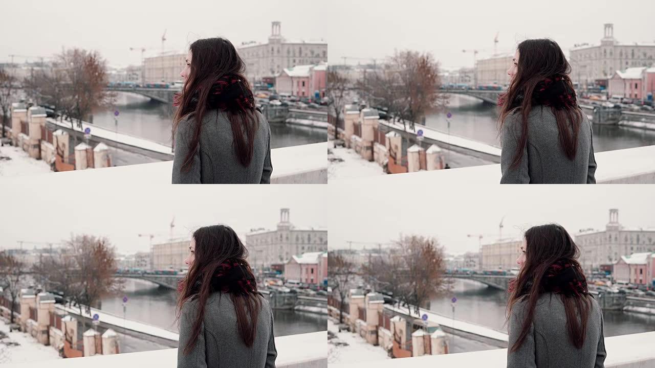 后视图。迷人的年轻黑发女孩站在桥上，看着冰雪覆盖的冬季小镇