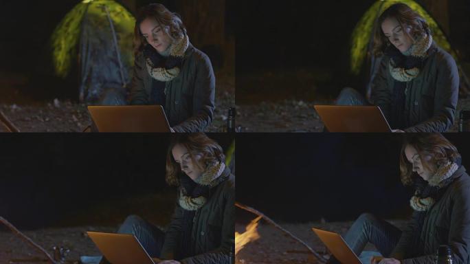 布鲁内特女孩在晚上的篝火旁使用笔记本电脑，背景是帐篷。