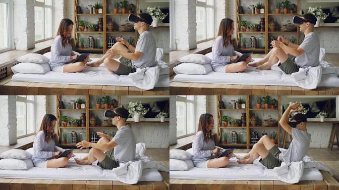 年轻夫妇玩得开心男人戴着虚拟现实眼镜，女人触摸平板电脑屏幕，他们坐在卧室的双人床上说笑。