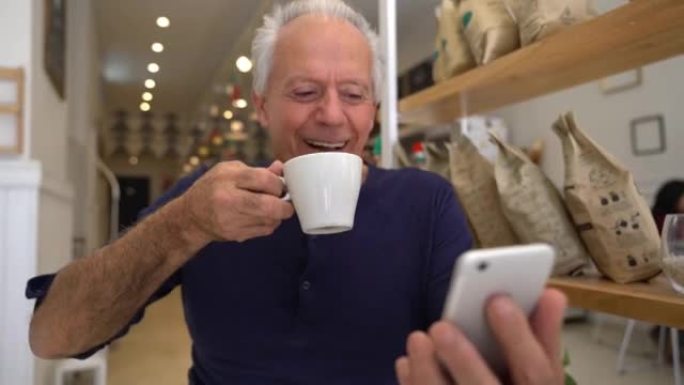资深男子在智能手机上观看视频，在面包店笑着喝咖啡