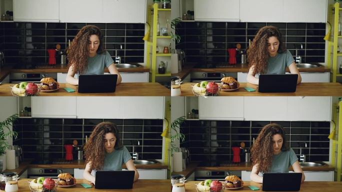年轻漂亮卷曲的白人妇女独自坐在漂亮宽敞的厨房里的餐桌旁工作，他们在笔记本电脑上听音乐、在笔记本电脑上