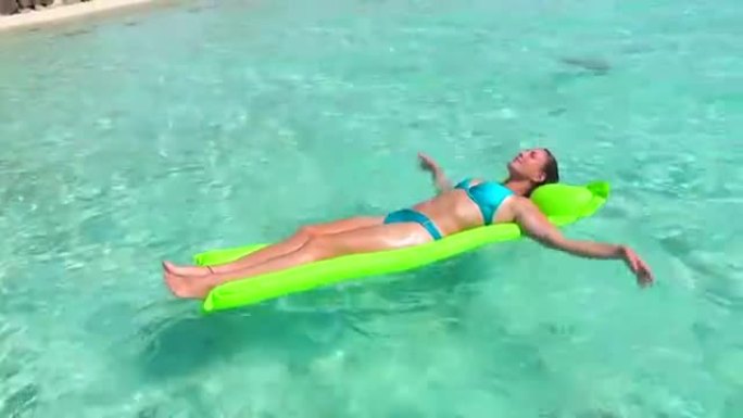 特写: 微笑的比基尼年轻女子在漂浮在热带岛屿清澈的海洋水面上的充气充气床垫上享受和放松