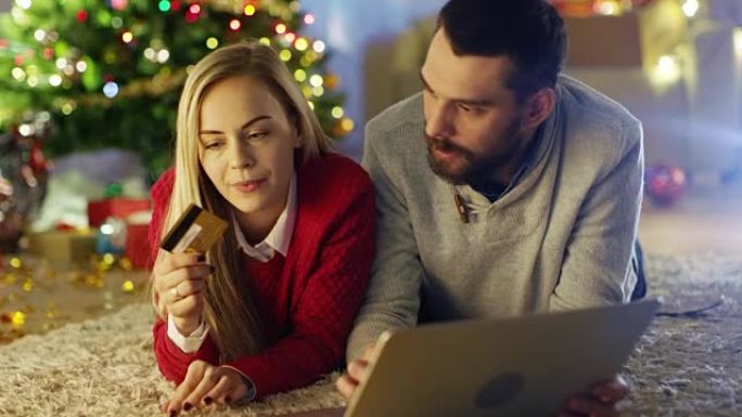 幸福的夫妻躺在圣诞树下的地毯上，女人拿着信用卡，男人在笔记本电脑上买礼物。