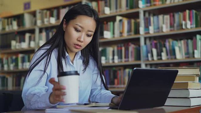 年轻的亚洲学生女孩坐在桌子旁，在大学图书馆里堆着书，用笔记本电脑工作，喝咖啡微笑着看着远处，准备考试