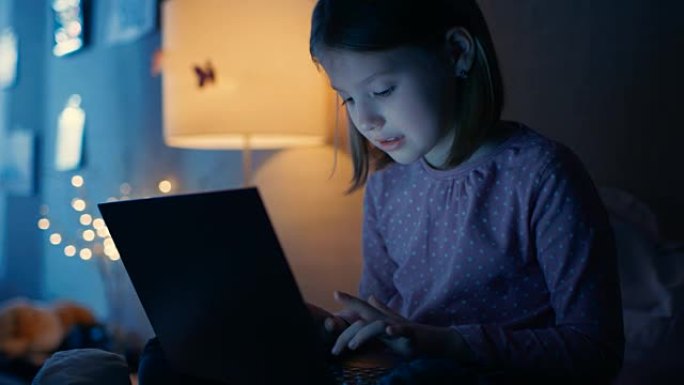 聪明的年轻女孩在深夜的卧室里坐在她的床上，用笔记本电脑打字一些有趣的东西。她的夜灯亮着。