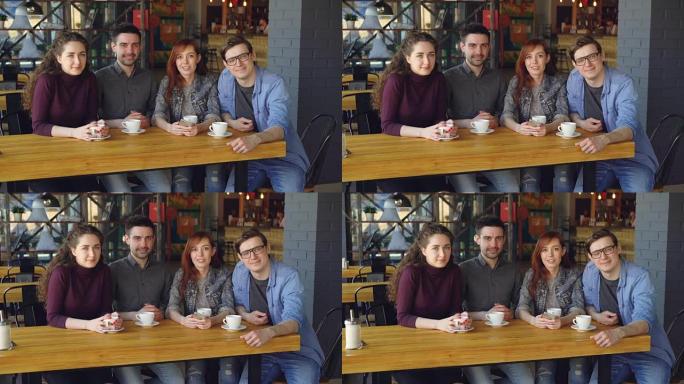 四个穿着便服的年轻人朋友的肖像坐在宽敞的咖啡馆里的桌子旁，拿着茶杯，看着相机。外出就餐和友谊概念。