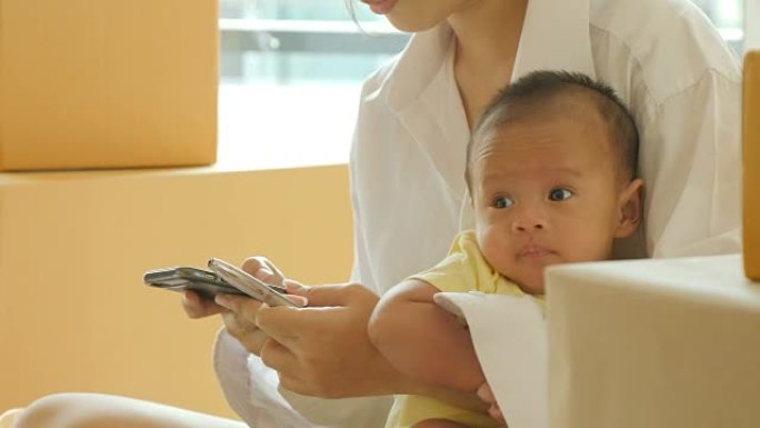 年轻的亚洲母亲抱着婴儿，并从智能手机上检查客户订单，并在线交付以准备在卧室包装。