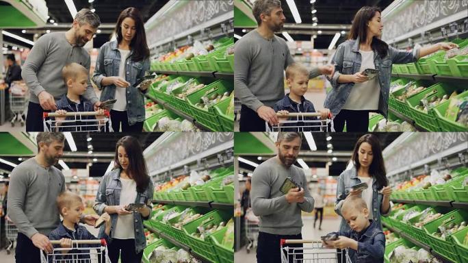 英俊的大胡子家伙，他迷人的妻子和可爱的孩子正在超市的托盘中选择蔬菜，说话，大笑并将产品放在手推车中。