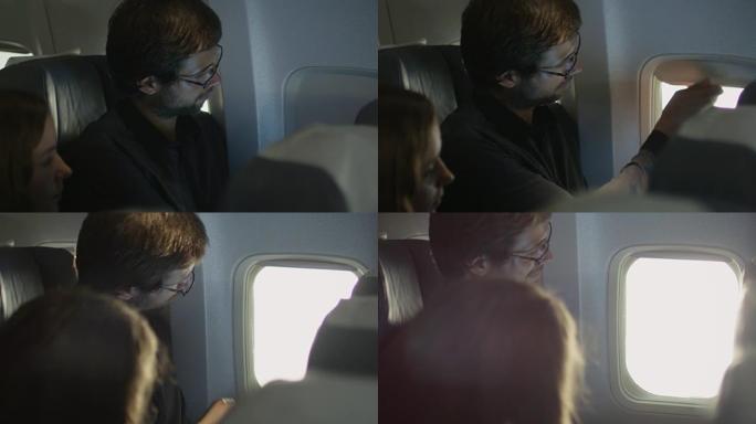 年轻人打开飞机上的窗帘，看着外面，而一个女人坐在他旁边。