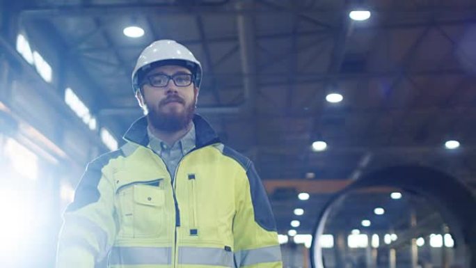戴着安全帽的工业工程师戴着安全帽挥手致意，走过各种金属加工工艺的重工业制造厂。