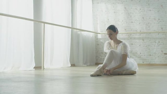 年轻漂亮的芭蕾舞演员穿着芭蕾舞短裙坐在木地板上，系着她的脚尖鞋。