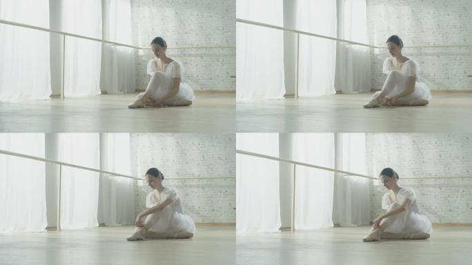 年轻漂亮的芭蕾舞演员穿着芭蕾舞短裙坐在木地板上，系着她的脚尖鞋。