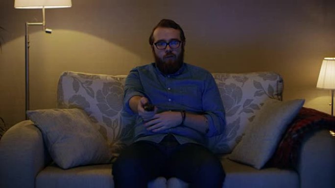 一个男人坐在客厅的沙发上，看电视，换频道的肖像。落地灯打开。
