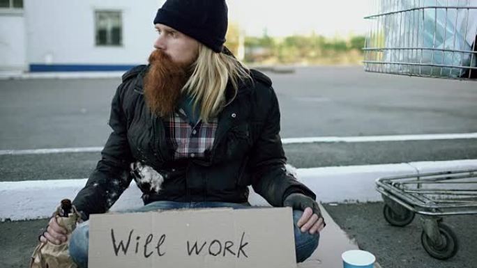 倾斜年轻的无家可归的人，用纸板坐在购物车附近，在寒冷的日子里喝酒