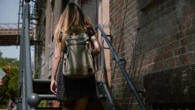 后视图的年轻女子背着双肩包和长发走上旧的金属楼梯进入红砖建筑缓慢的动作