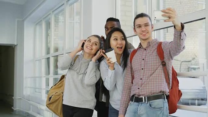 站在大学走廊上的一群多种族学生在智能手机相机上自拍。手持电话和朋友的时髦家伙摆出积极的姿势