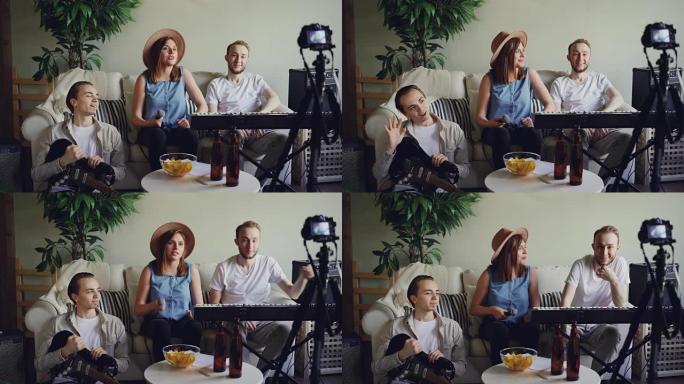 快乐的朋友音乐家正在使用相机为他们的互联网博客录制视频。年轻人坐在家庭工作室的沙发上说话，打手势和大