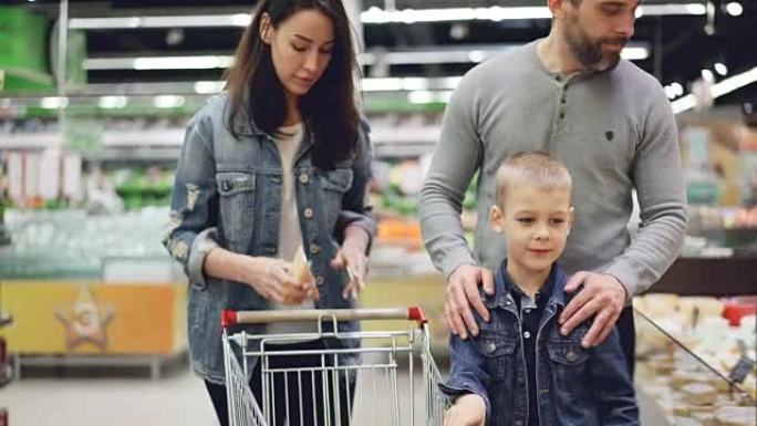 母亲，父亲和儿子幸福的家庭正在超市的乳制品部门选择奶酪，他们从冰箱中取出碎片并闻起来，然后放入购物车